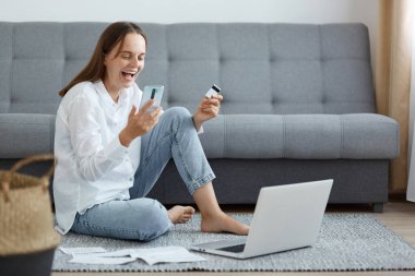 Kanepenin yanında oturan, dizüstü bilgisayar ve telefon kullanan, kredi kartı tutan, piyangoyu kazanan ve heyecanını dile getiren, beyaz gömlek ve kot giyen mutlu ve mutlu bir kadın..