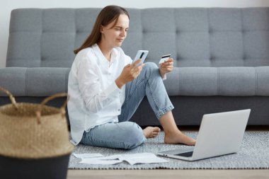 Beyaz gömlekli ve kot pantolonlu çekici bir kadın görüntüsü kanepenin yanında oturuyor, dizüstü bilgisayar ve telefon kullanıyor, kredi kartı tutuyor, online ödeme verilerini kontrol ediyor..