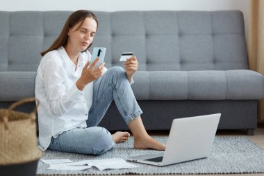 Beyaz gömlek ve kot pantolon giyen genç bir kadının, koltuğun yanında otururken, cep telefonu kullanırken, kredi kartı tutarken, online ödeme verilerini incelerken.