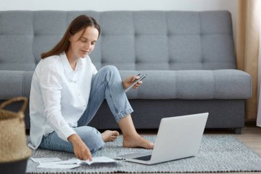 Beyaz gömlek ve kot pantolon giyen beyaz kadın portresi kanepenin yanında oturuyor, dizüstü bilgisayar kullanıyor, kredi kartı ve cep telefonu kullanıyor, kağıt dökümanlara bakıyor..