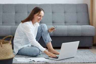 Mutlu, gülümseyen, beyaz gömlek ve kot giyen, kanepenin yanında oturan, dizüstü bilgisayar kullanan, kredi kartı ve cep telefonu kullanan, olumlu duygularını ifade eden, online ödeme yapan çekici bir kadın..