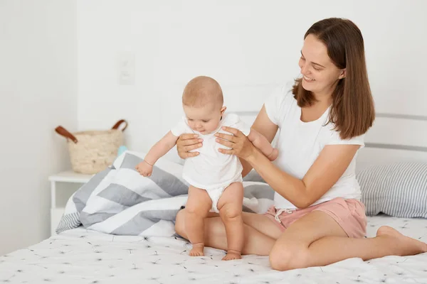 室内拍摄的黑发女子身穿白色T恤 与女婴坐在床上玩耍 小孩在妈妈的帮助下站立 快乐的一家人在一起度过时光 — 图库照片