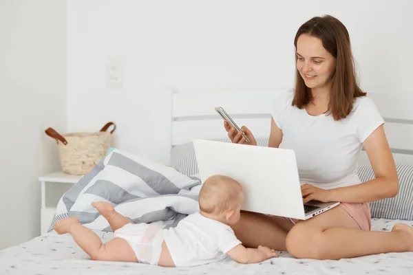 漂亮漂亮的女人与女婴一起躺在床上 上网工作 拿着笔记本电脑和手机 把产假和上网工作结合起来 摆出一副很不错的姿势在卧室里 — 图库照片