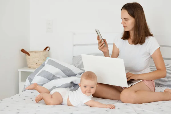 白种人妇女与女婴躺在床上上网工作 两腿牵着笔记本电脑 手机拨号 打电话给伴侣或保姆的横向照片 — 图库照片
