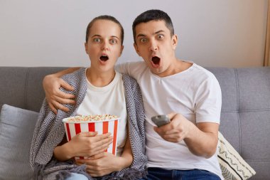 Kanepede patlamış mısır ve uzaktan kumandayla oturan, korku filmi izleyen, ağzı açık kameraya bakan, şok olmuş ve korkmuş genç bir çiftin çekimleri..