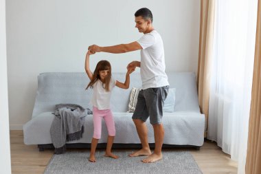 Beyaz bir tişört giyen, kızıyla evde oynayan, mutlu bir şekilde dans eden, birlikte mutlu vakit geçiren pozitif bir adamın uzun bir portresi..
