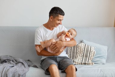Beyaz bir tişört giymiş, genç ve yakışıklı bir adamın kanepede elinde bebekle oturup kızına sevgiyle bakışının yatay görüntüsü..