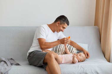 Beyaz tişört giyen esmer esmer adamın portresi. Kanepede oturuyor ve küçük bebekle oynuyor. Sevimli kızına oyuncağını gösteriyor..