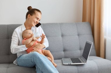 Beyaz gömlek ve kot pantolon giyen gülümseyen bir kadının yan görünüm portresi. Küçük tatlı çocuğuyla koltukta oturuyor. Not defteri kullanıyor ve telefonla konuşuyor..