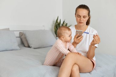 Siyah saçlı, beyaz tişörtlü ve şortlu bir kadın bebeğiyle aynı yatakta oturuyor ve hamilelik testi negatif sonuç veriyor, biriyle konuşmak için numarayı çeviriyor, hayal kırıklığı yaşıyor..