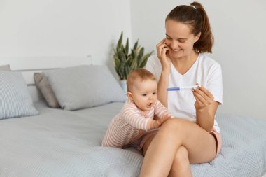 Beyaz tişört ve şort giyen mutlu bir kadının bebek kızıyla yatakta oturup hamilelik testi yapıp kocasını iyi haberler vermeye çağırması..