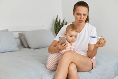 Beyaz tişört ve şort giyen üzgün bir kadın bebeğiyle aynı yatakta oturuyor ve hamilelik testi pozitif sonuç veriyor..