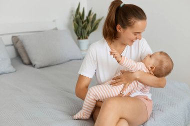 Beyaz t-shirt ve şort giyen mutlu çekici bir kadının portresi. Bebek kızıyla yatakta oturuyor. Annesi çocuğuyla oynuyor..