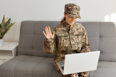 Kapalı alanda gülümseyen tatmin olmuş kadın asker, kamuflaj üniforması ve şapka giyip kanepede oturuyor ve laptopta çalışıyor, video görüşmesi yapıyor, el sallıyor, merhaba diyor..