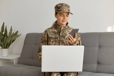 Kafkas askeri kadının kamuflaj üniforması ve şapkası ile kanepeye oturup dizüstü bilgisayarda çalışırken, akıllı telefon kullanarak, cihaz ekranına bakarken yatay görüntüsü..