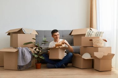 Beyaz tişörtlü esmer bir adamın karton kutularla çevrili kanepenin yanında oturduğu yatay poziyonu. Taşındıktan sonra şahsi eşyalarını üst üste yığıyor..