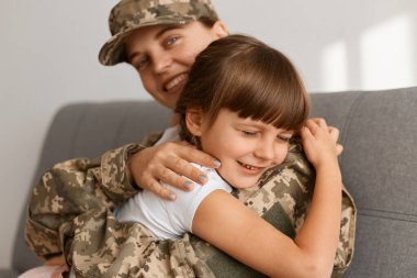Son derece mutlu, koyu renk saçlı bir kızın yakın portresi. Annesinin askerine sarılıyor. Kamuflaj üniforması giyen ve kızıyla poz veren bir kadın..