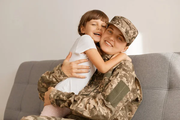 Indoor shot of delighted extremely happy military woman wearing camouflage uniform and cap posing with her daughter at home while sitting on sofa, hugging, mother come back home from war.