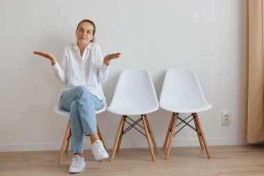 Kafası karışmış, çaresiz, beyaz gömlekli, kot pantolonlu, duvara yaslanmış, ellerini yana açmış, omuz silkmiş, cevap alamayan, kameraya bakan bir kadın..