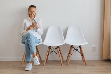 Beyaz gömlekli, kot pantolonlu, sırasını beklerken cep telefonu kullanan beyaz bir kadının kapalı çekimleri..