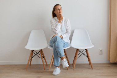 Beyaz gömlek ve kot pantolon giyen, duvara yaslanmış kadın bakışları ve çenesini kaldırıp önemli şeyler düşünürken..