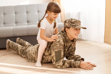 Gülümseyen beyaz kadın askerin tam boy portresi. Kamuflaj üniforması ve şapkası giyen, kızıyla birlikte vakit geçirmekten mutlu olan kadın asker..