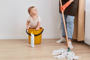 İç mekanda, bebek bebek tulumlu, sarı kovada oturan, yüzünü belli etmeyen, kot pantolon ve önlük giyen, çiçekleri yıkayan bir bebek resmi..