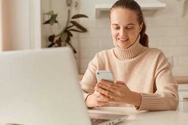 Mutfakta bej kazak giyen, elinde akıllı bir telefonla sosyal ağları kontrol eden, fotoğraf izleyen ve gülümseyen çekici, koyu renk saçlı bir kadın resmi..