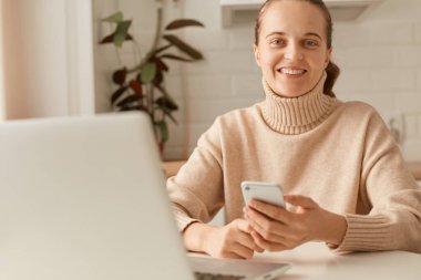 Tatmin olmuş, bej kazak giyen, dişlek gülüşlü kameraya bakan, cep telefonunu elinde tutan, olumlu duygularını ifade eden bir kadının görüntüsü..