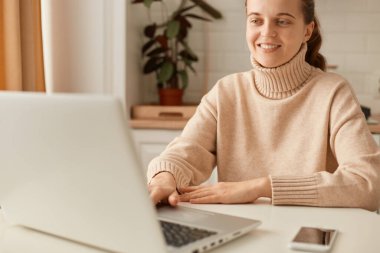 Mutfakta bej kazak giyen ve bilgisayar başında çalışan mutlu bir kadın. Dişlek gülümsemeli dizüstü bilgisayara bakıyor. İnternette çalışan bir kadın..