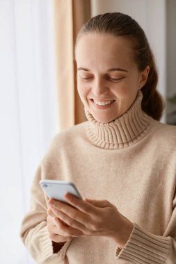 Sevimli, çekici, pozitif bir kadının elinde cep telefonuyla pencerenin yanında dikilen bej kazak giyip neşeli iyimser yüz ifadesi olan bir cihaz kullanması ve komik videolar izlemesi..