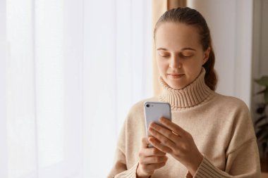 Kapalı alanda gülümseyen, bej kazak giyen, elinde cep telefonuyla pencerenin yanında duran, çevrimiçi kaydıran, daktilo eden, sosyal ağları kontrol eden ya da mesajlara cevap veren çekici bir kadın resmi..