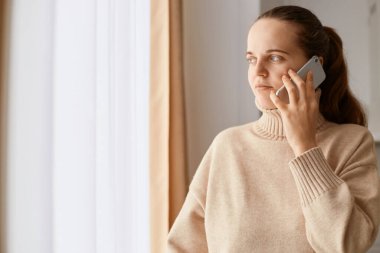 Koyu renk saçlı, at kuyruklu, bej kazaklı, pencerenin yanında poz veren, akıllı telefonla konuşan ve yüzünü çeviren, sakin bir yüz ifadesi olan bir kadın..