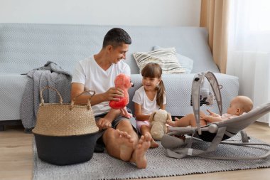 Beyaz tişörtlü esmer bir adamın, kızlarıyla yerde otururken, babasının sallanan kızına yumuşak oyuncağını gösterirken, bebek ve anaokulu çocuklarıyla kanepenin yanında poz verirken..