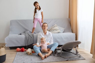 Beyaz gömlek ve kot pantolon giymiş üzgün bir kadının uzun bir portresi. Kızlarıyla yerde oturuyor. Bunalımda ve yorgun ve gürültücü çocuklara bakıyor..