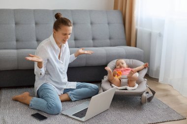 Çaresiz bir kadının portresi, beyaz gömlek ve kot pantolon giymiş koltuğun yanında oturuyor, bebek sallanan sandalyede oturuyor ve not defterine video çağrısı yapıyor, omuzlarını silkiyor, emin değilim, cevap vermiyor..