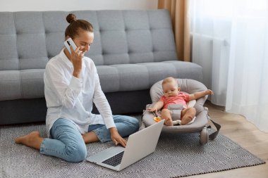 Bun saçlı, beyaz gömlekli ve kot pantolonlu bir kadının sallanan sandalyesinde bebek otururken yerde oturması, cep telefonuyla konuşması ve not defteriyle yazması..