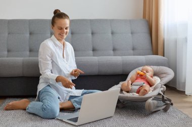 Beyaz gömlek ve kot pantolon giyen, sallanan sandalyede bebeği olan ve her iki işaret parmağıyla dizüstü bilgisayarı gösteren güzel, iyimser bir kadının tam boy portresi..