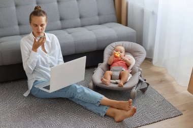 Koyu renk saçlı, beyaz gömlekli ve kot pantolonlu bir kadının sallanan sandalyede bebeği olan koltuğun yanında oturması, ses asistanı kullanması, mesaj kaydetmesi, küçük kızına bakması..