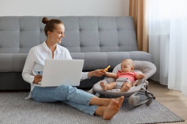 Beyaz gömlekli, kot pantolonlu, sallanan koltuğun yanında bebek koltuğunda oturan, annesinin bilgisayar başında çalışırken küçük kızıyla oynaştığı kapalı mekan fotoğrafı..