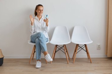 Beyaz gömlek ve kot pantolon giyen, kuyrukta oturan, canlı yayın yapan ya da video çağrısı yapan, elini sallayarak merhaba diyen çekici, koyu renk saçlı bir kadın..