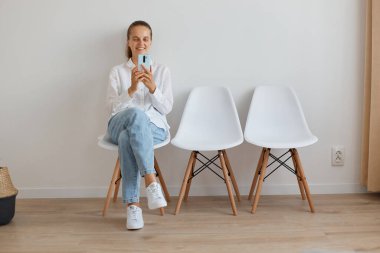 Beyaz gömlekli ve kot pantolonlu bir kadının uzun bir portresi kuyrukta otururken ve telefon kullanırken ya da video araması yaparken mutlu bir şekilde gülümserken..