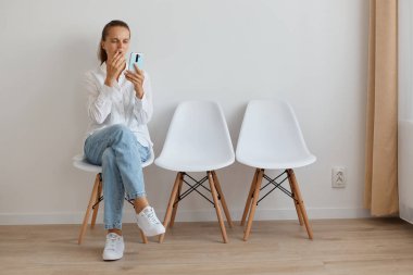 Beyaz gömlek ve kot pantolon giyen, kuyrukta oturan, cep telefonu tutan ve ağzı palmiyeyle kaplanmış bir kadının tam boy portresi..