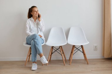 Beyaz gömlek ve kot pantolon giyen beyaz bir kadının portresi kuyrukta oturuyor, telefonla konuşuyor, gülümsüyor ve olumlu duygular ifade ediyor..