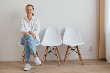 Beyaz gömlek ve kot pantolon giyen mutlu bir kadının portresi kuyrukta oturuyor, ellerini bacaklarının üzerinde tutuyor, iyimser bir ifadeyle kameraya bakıyor..