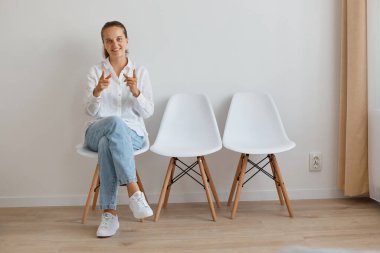 Beyaz gömlek ve kot pantolon giyen, kuyrukta oturan, kameraya bakıp işaret eden mutlu ve mutlu genç bir kadının tam boy portresi..