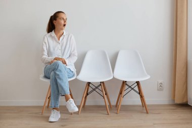 Beyaz gömlek ve kot pantolon giyen beyaz bir kadın heyecanlı kuyrukta oturuyor, ağzı açık bakıyor, şok ve şaşkınlık ifade ediyor.