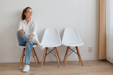 Atkuyruğunda oturan beyaz gömlekli, kot pantolonlu, kendine güvenen güzel bir kadın. Sakin görünüyor, gülümseyerek yüzünü çeviriyor, olumlu duygular ifade ediyor..