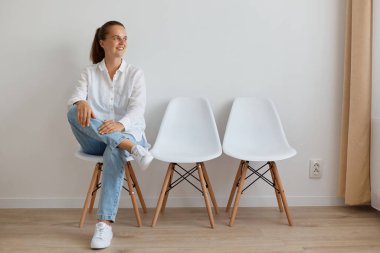 Beyaz gömlek ve kot pantolon giyen güzel, gülümseyen bir kadının uzun bir portresi kuyrukta oturuyor, iyimserlikle başka yere bakıyor, bekliyor..