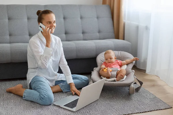 Beyaz gömlekli, kot pantolonlu, sallanan koltuğun yanında oturan, genç bir annenin telefonla konuşan ve dizüstü bilgisayarla çalışan oldukça koyu renk saçlı bir kadının portresi..